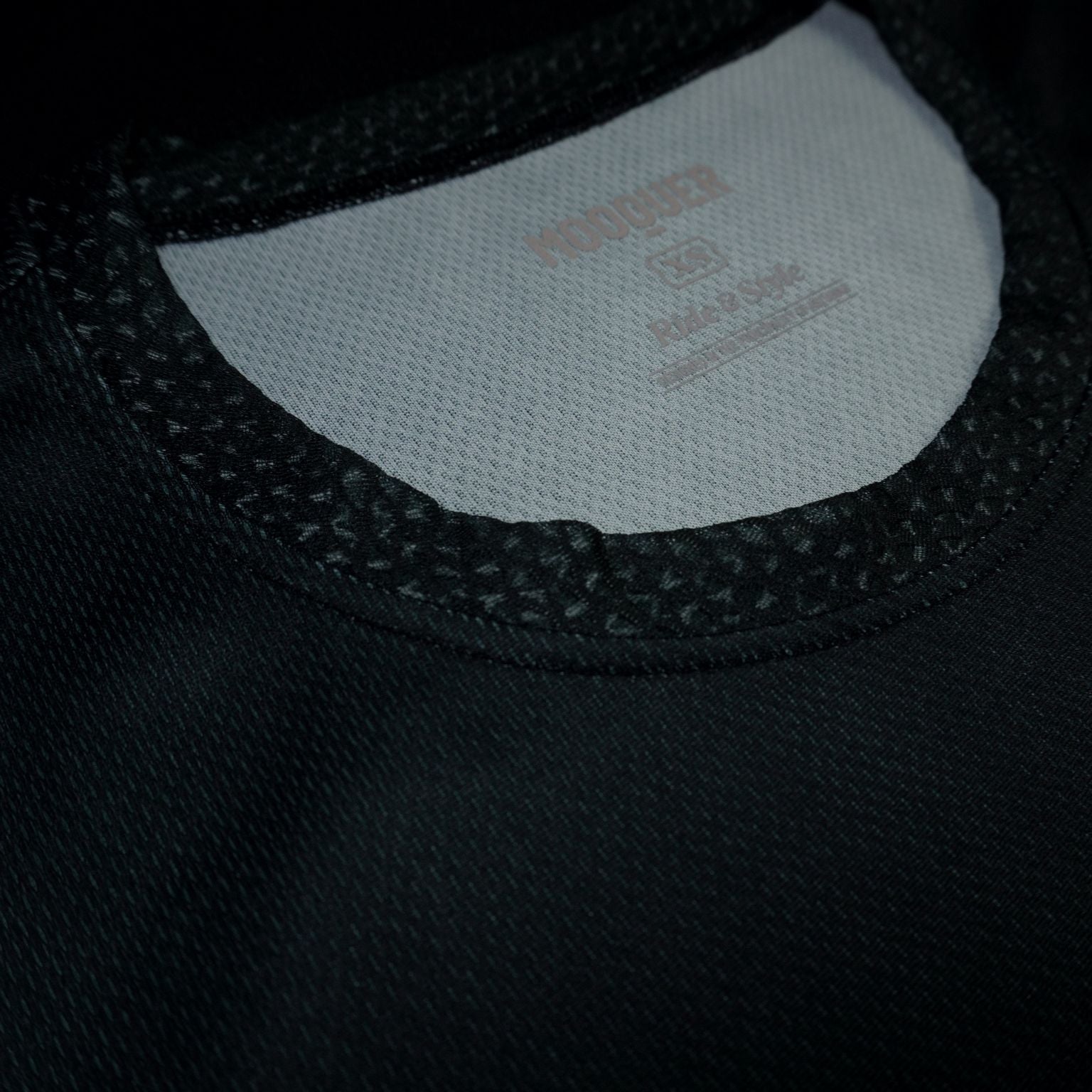 Detalle del bordado MOOQUER en hilo gris claro sobre tejido negro de la camiseta lifestyle